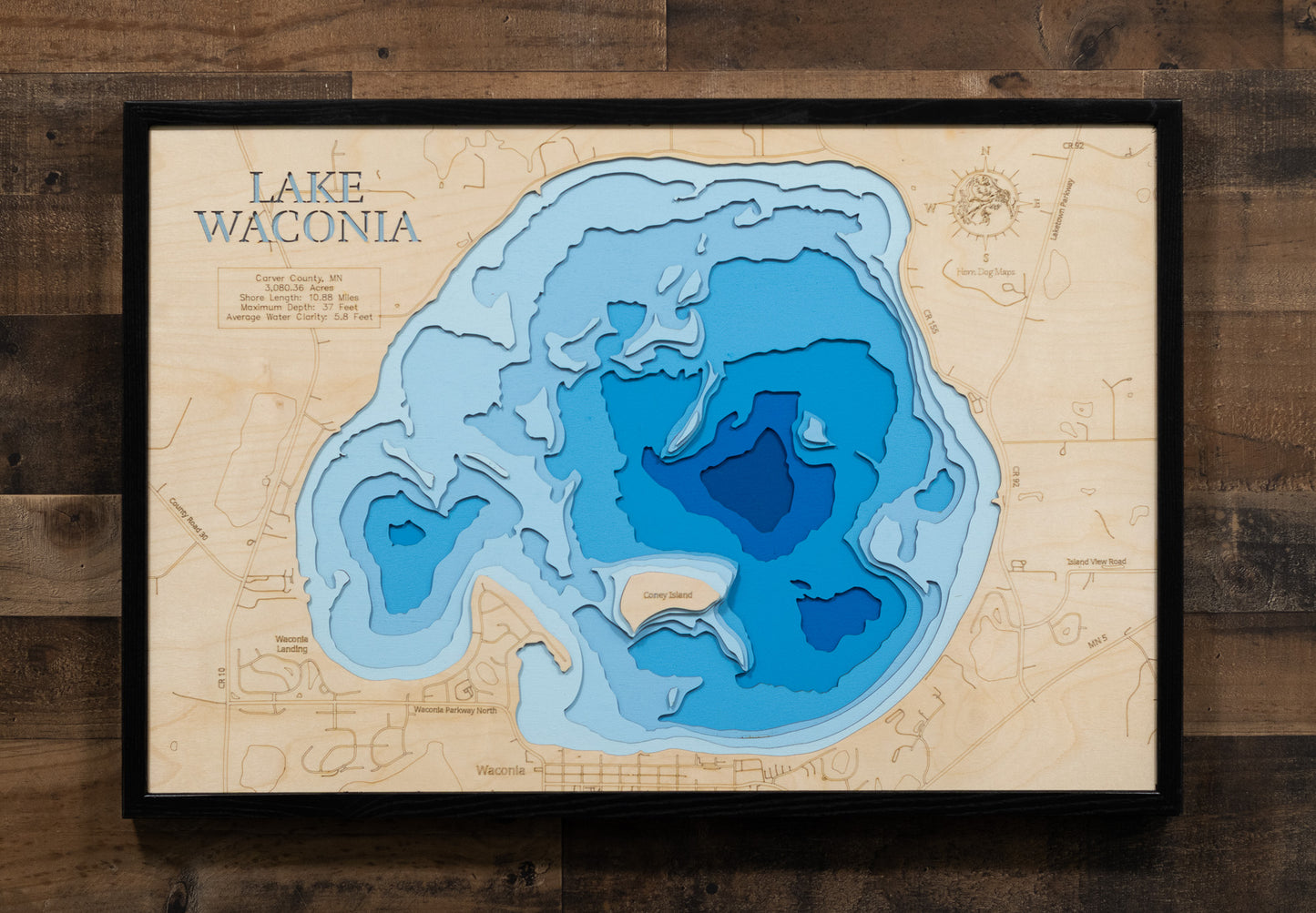 A laser-cut, layered wooden map of Lake Waconia in Carver County, Minnesota. The distinct shape of this natural lake, with its 10.88-mile shoreline and prominent islands, is clearly visible within the suburban and agricultural landscape of the Minneapolis metropolitan area. The varying depths of the lake, reaching a maximum of 37 feet, are represented by different layers of wood, illustrating its impressive underwater topography.