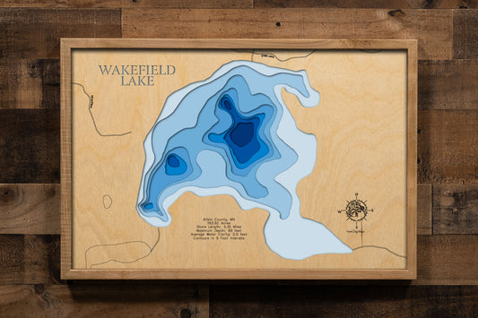 A custom-engraved, laser-cut wooden map of Wakefield Lake in Aitkin County, Minnesota. The map features precise bathymetric details with engraved contour lines at 9-foot intervals, capturing the lake's impressive depth and unique underwater topography. The water is represented by a deep blue stain, contrasting with the natural grain of the wood. The map includes elegant etchings of key lake data such as area, shore length, and maximum depth, providing both a beautiful piece of art and a detailed, informativ