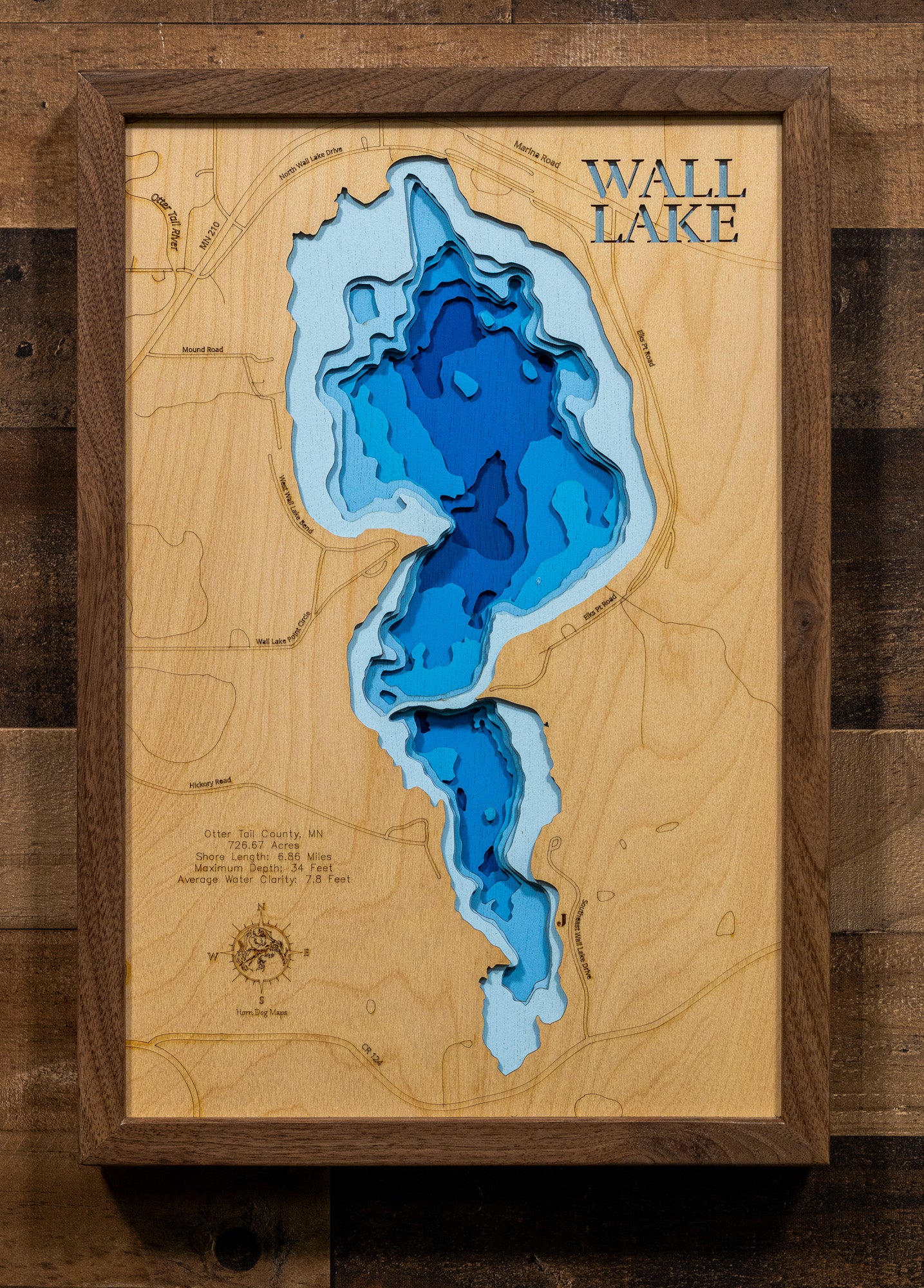 A framed 3D wooden wall art piece depicting Wall Lake, with layered blue and light blue wood sections forming the lake shape over a natural wood map background. The words 'WALL LAKE' appear in the top right, and the piece is mounted against a dark wood plank wall.