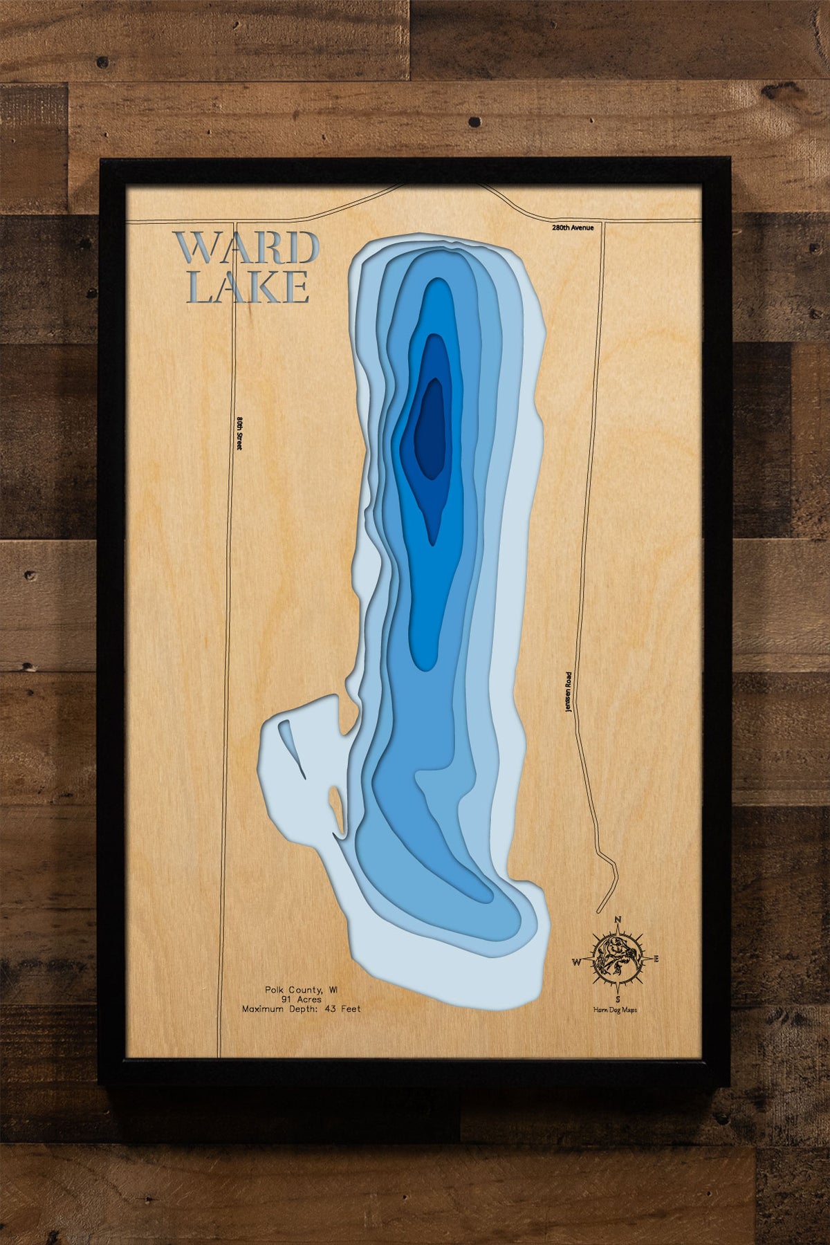 A precision laser-cut, multi-layered wooden map of Ward Lake in Polk County, Wisconsin, a 91-acre seepage lake in the northern part of the county near Luck and Frederic with a notable maximum depth of 43 feet. The natural wood top layer captures the lake's intimate shoreline in fine detail, while a light-to-dark blue gradient conveys its depth and cold-water character. A handcrafted statement piece for any Wisconsin lake cabin, home, or outdoor enthusiast's wall.