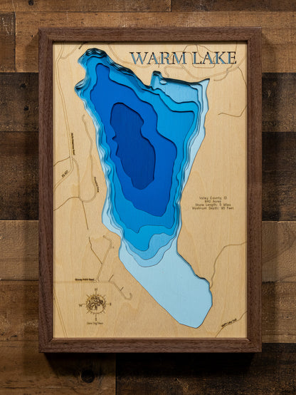 A laser-cut, layered wooden map of Warm Lake in Valley County, Idaho. The distinct oval shape of this high-mountain lake, with its 5-mile shoreline, is clearly visible within the rugged landscape of the Payette National Forest. The varying depths of the lake, reaching a maximum of 95 feet, are represented by different layers of wood, illustrating its impressive underwater topography.
