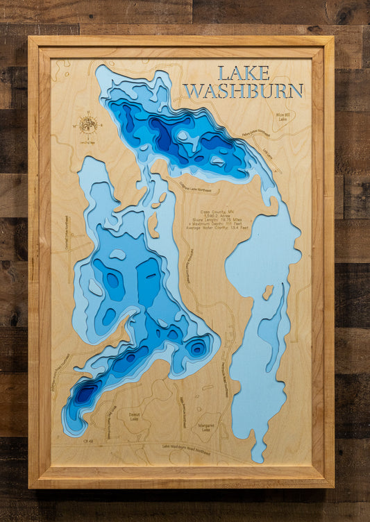 A framed 3D wooden topographic map of Lake Washburn, featuring layered blue wood sections representing different depths on a light natural wood background, enclosed in a light wood frame and mounted on a dark wood plank wall.