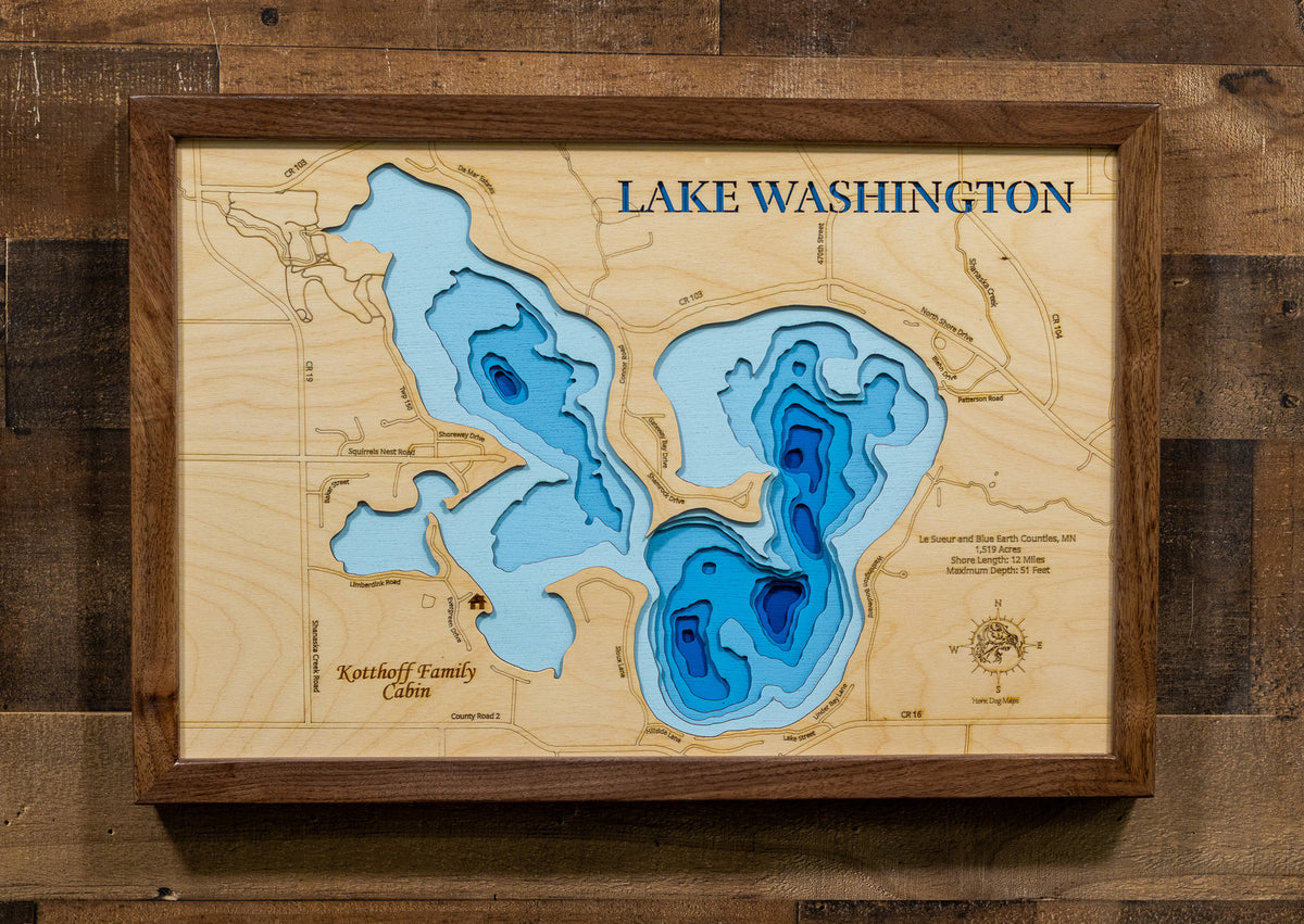 A framed wooden wall art piece depicting Lake Washington with layered blue depth contours and engraved text, mounted on a rustic wood plank wall.