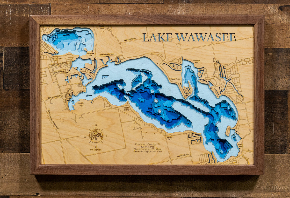 A framed 3D wooden depth map of Lake Wawasee, featuring layered blue and light blue wood sections with engraved details and the text 'LAKE WAWASEE' at the top, mounted on a rustic dark wood wall.