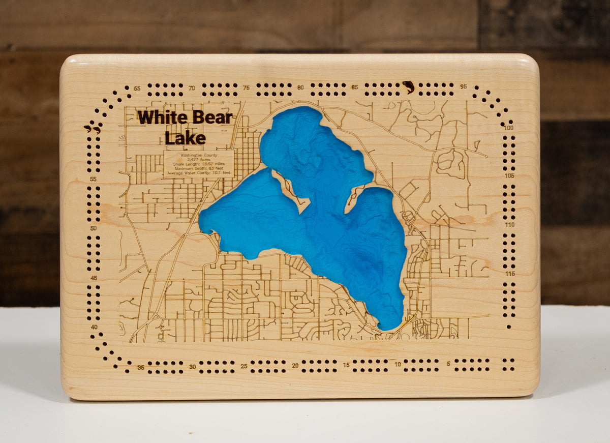 A rectangular wooden plaque featuring a laser-engraved map of White Bear Lake, with a raised blue lake shape in the center and black engraved text reading 'White Bear Lake' on a light wood background.