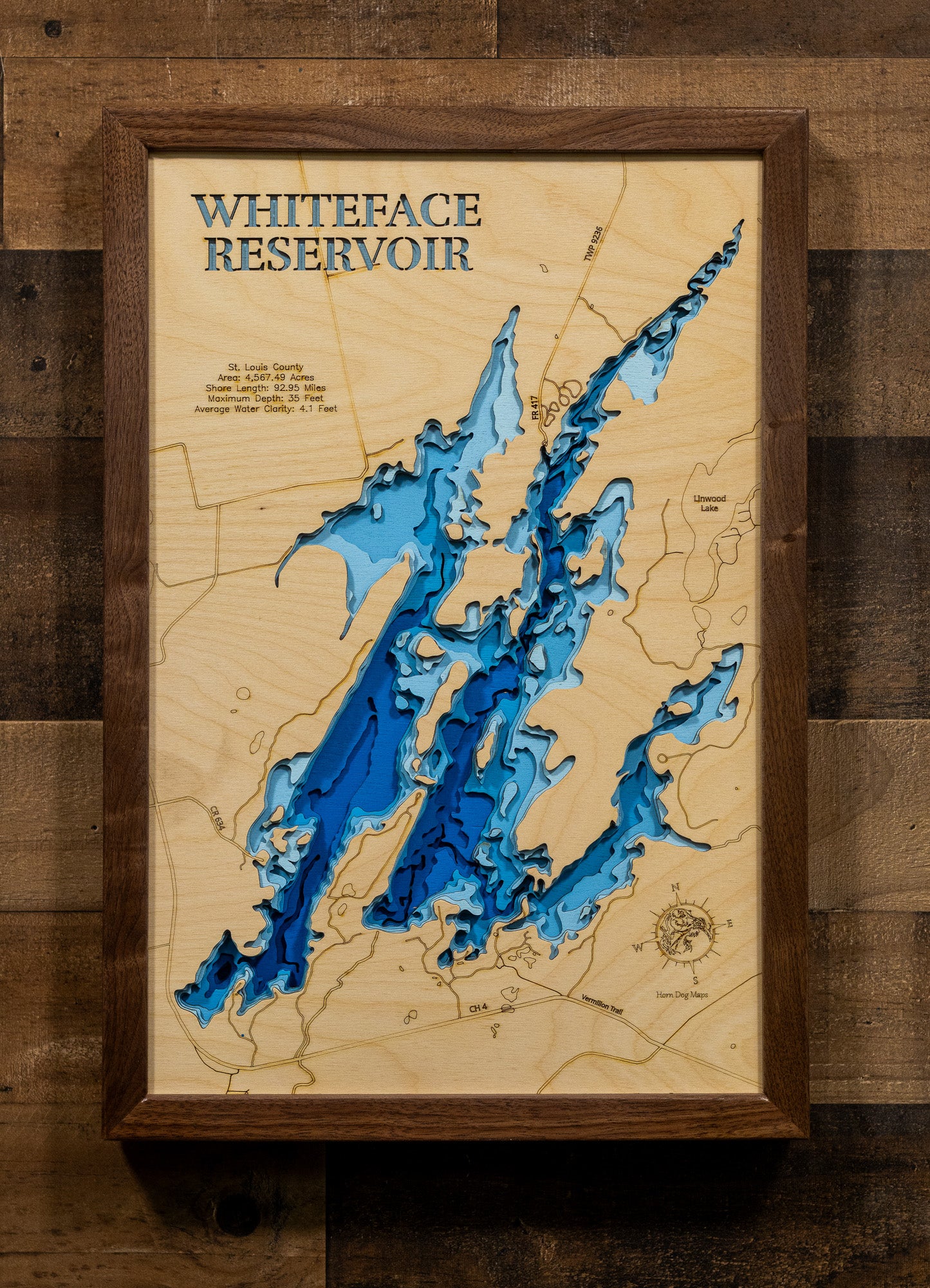 A framed 3D wooden topographic map of Whiteface Reservoir, featuring layered wood cutouts in shades of blue and brown over a light wood background, mounted on a rustic dark wood plank wall.
