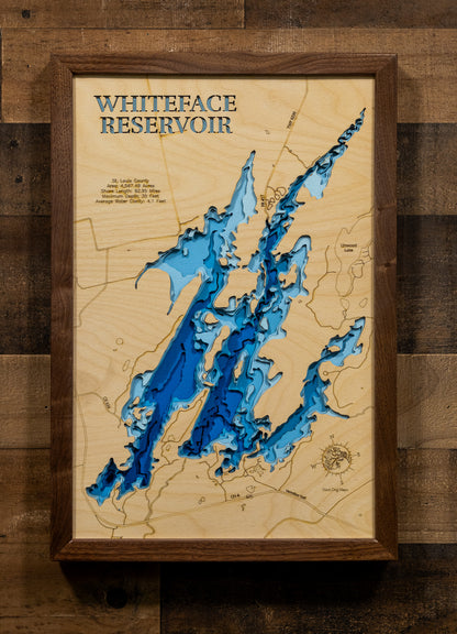 A framed 3D wooden topographic map of Whiteface Reservoir, featuring layered wood cutouts in shades of blue and brown over a light wood background, mounted on a rustic dark wood plank wall.