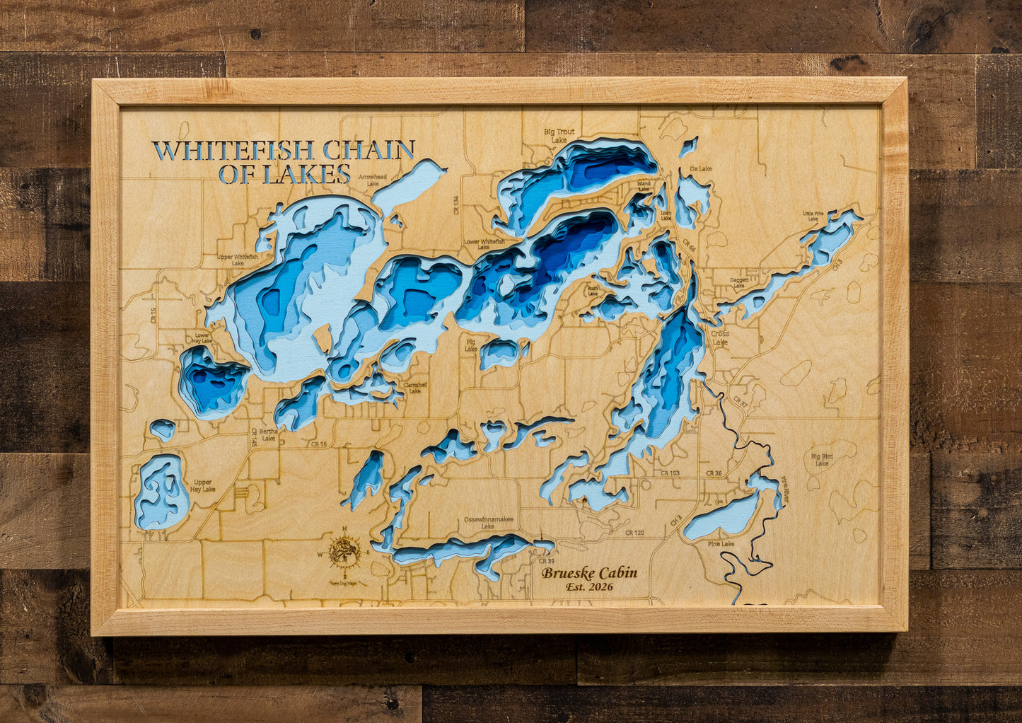 A framed wooden map of Whitefish Chain of Lakes, featuring layered blue and light blue wood cutouts representing water depths, mounted on a dark wood plank wall.