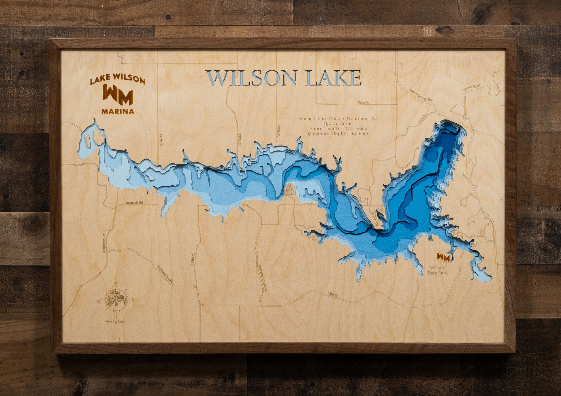 A laser-cut, layered wooden map of Wilson Lake, located in Russell and Lincoln Counties, Kansas. The shape of the reservoir, nestled within the Smoky Hills, is clearly visible within the central Kansas landscape. The varying depths of the lake, reaching a maximum of 65 feet, are represented by different layers of wood.