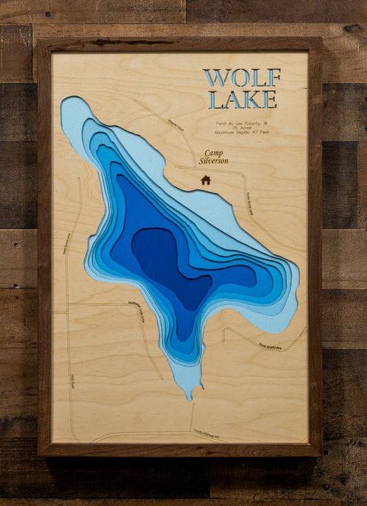 A framed wooden topographic map art print of Wolf Lake, featuring layered blue contour lines on a light wood background, mounted on a dark rustic wood plank wall.