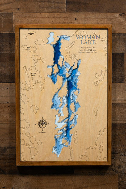 A magnificently detailed, laser-cut, multi-layered wooden topographical map of Woman Lake in Kenora District, Ontario, showcasing the expansive 4,771-acre wilderness lake with an incredible 68 miles of irregular rocky shoreline. The natural wood top layer frames the Woman River inlet, countless hidden bays and rocky points, and connections to the Woman River chain of lakes, while graduated blue layers create a stunning visual gradient from shallow rocky areas to the impressive 100-foot maximum depth.